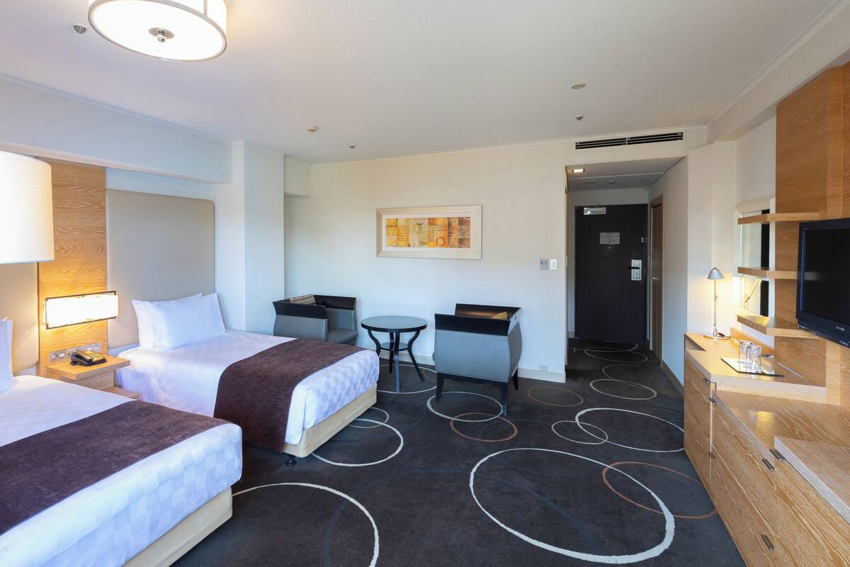 A hotel room with two beds, a desk, a chair, a TV, and a small table.