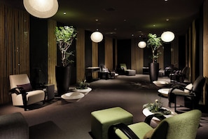 A modern lounge area with multiple seating options, including sofas and chairs, arranged around a central coffee table. There are large potted plants and hanging spherical light fixtures.
