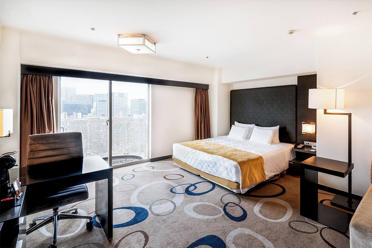 A hotel room with a large bed, a desk, a chair, and a view of the city.