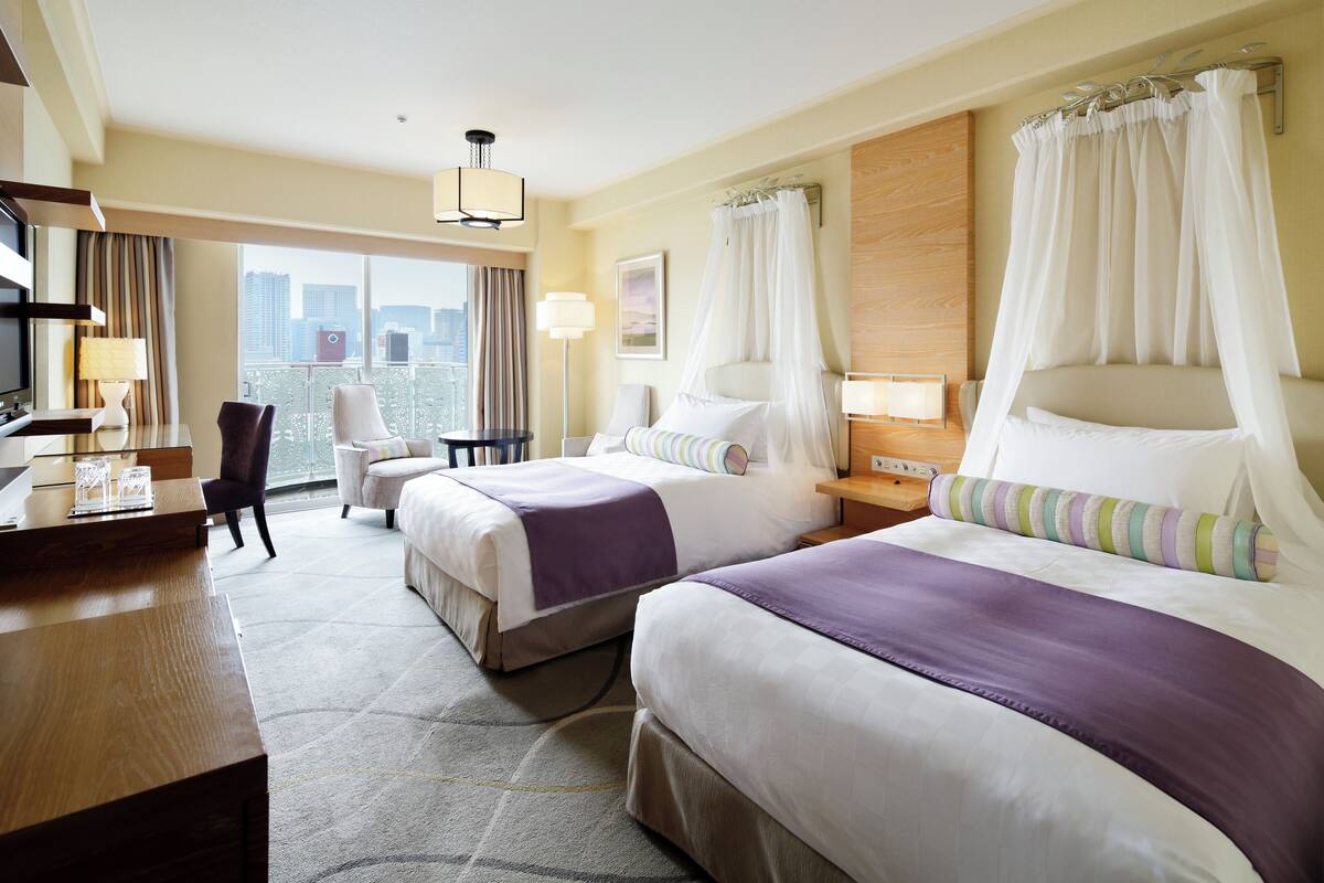 A hotel room with two beds, a desk, a chair, and a view of the city.