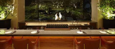 A modern bar area with a long counter, multiple wine glasses, and two large potted plants.