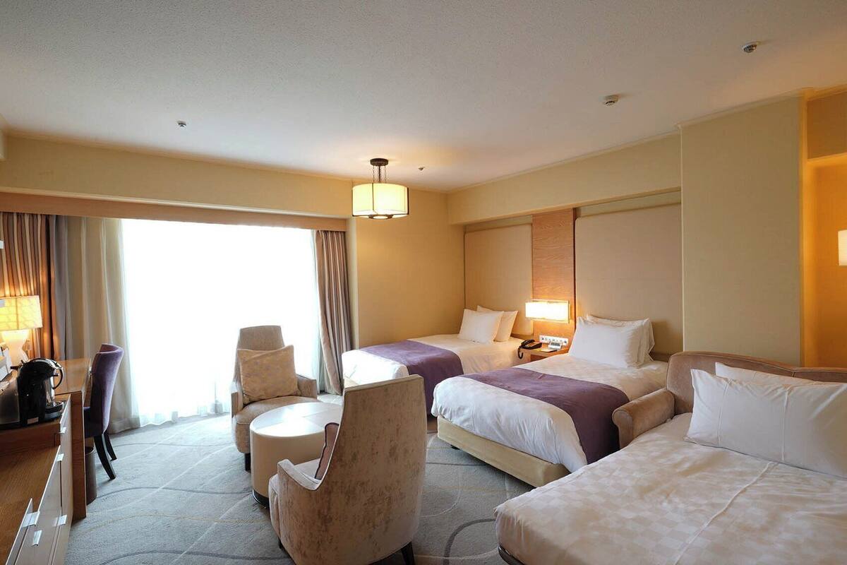 A hotel room with two beds, a sofa, a chair, a coffee machine, and a desk with a lamp.