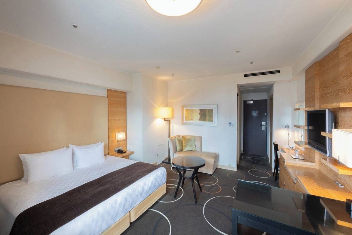 A hotel room with a bed, a desk, a chair, a lamp, and a television.