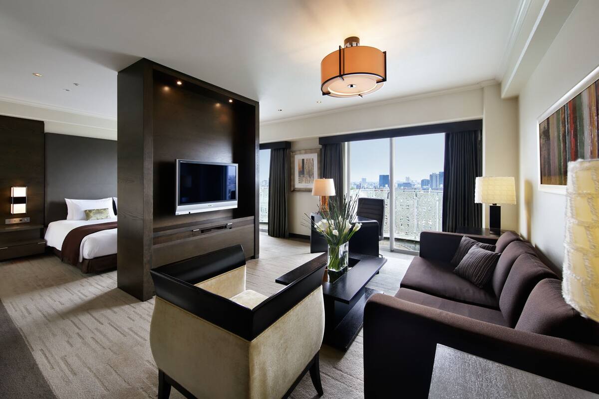 A modern hotel room with a large bed, a flat-screen TV, a sofa, a coffee table, and a city view.