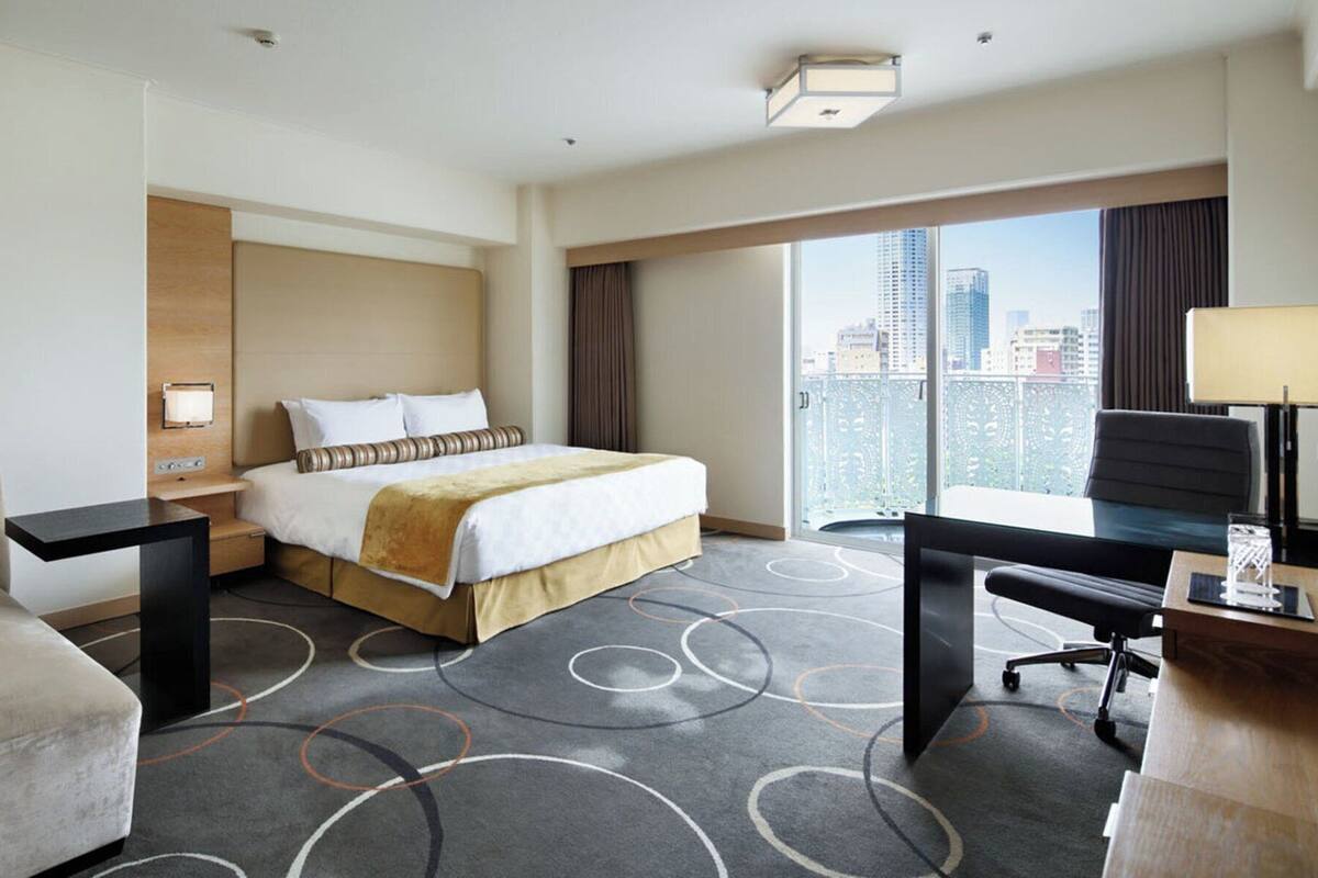 A hotel room with a large bed, a desk, a chair, a sofa, and a view of the city.