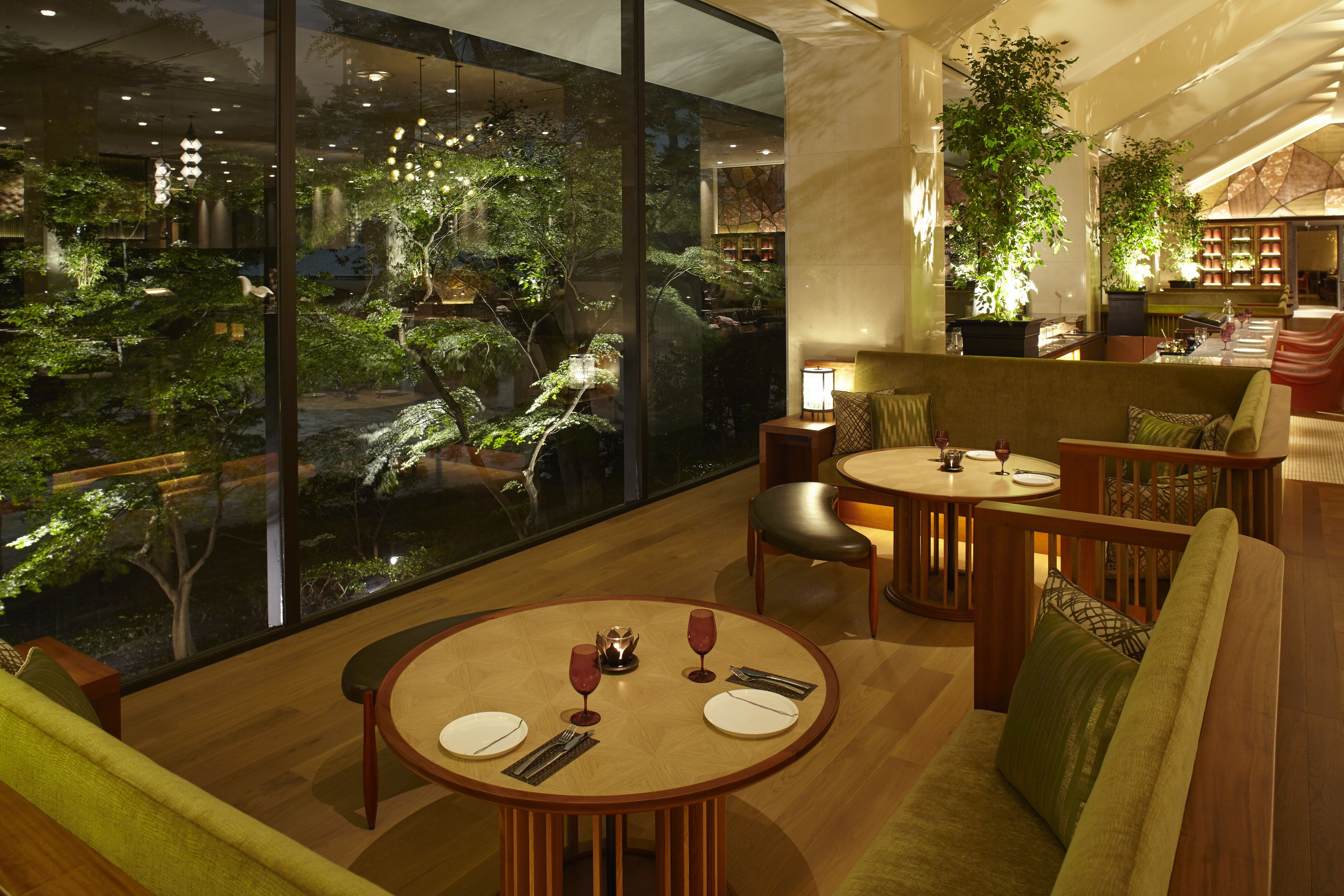 A modern dining area with a round table set for two, a sofa, and a view of a lush indoor garden through large windows.