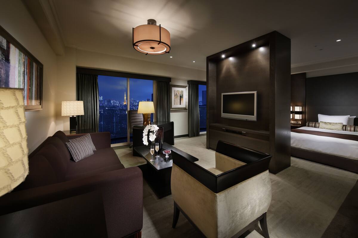A modern hotel room with a large bed, a TV mounted on the wall, a seating area with a sofa and armchair, a dining table with a vase and candles, and a large window offering a city view.