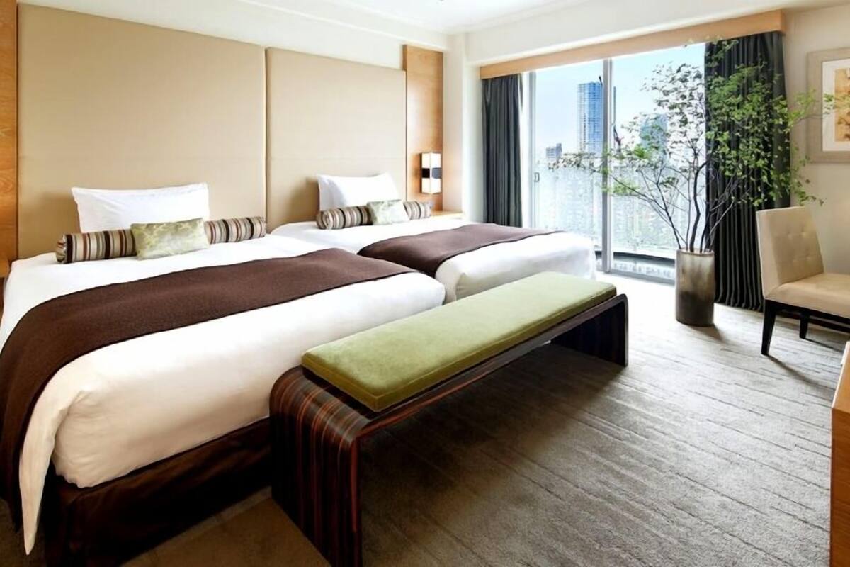 A hotel room with two beds, a bench, a chair, a desk, a window with a view of buildings, and a plant.