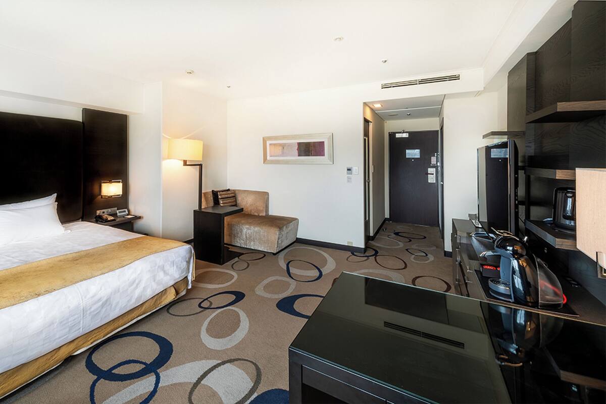 A hotel room with a large bed, a desk with a coffee machine, a chair, and a door leading to another room.