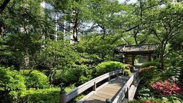 A wooden bridge in a lush garden with dense greenery and a traditional building in the background.
