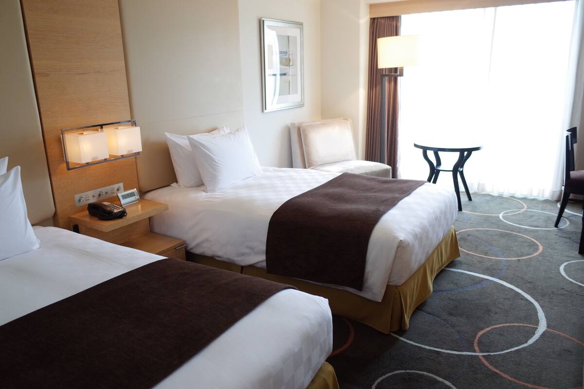 A hotel room with two beds, a bedside table with a lamp, a chair, and a view of the outdoors.