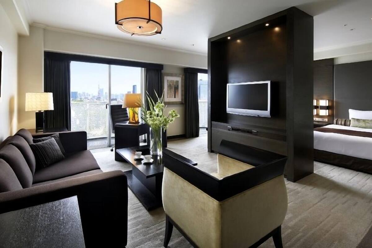 A modern hotel room with a sofa, a desk, a TV, a bed, and a city view.