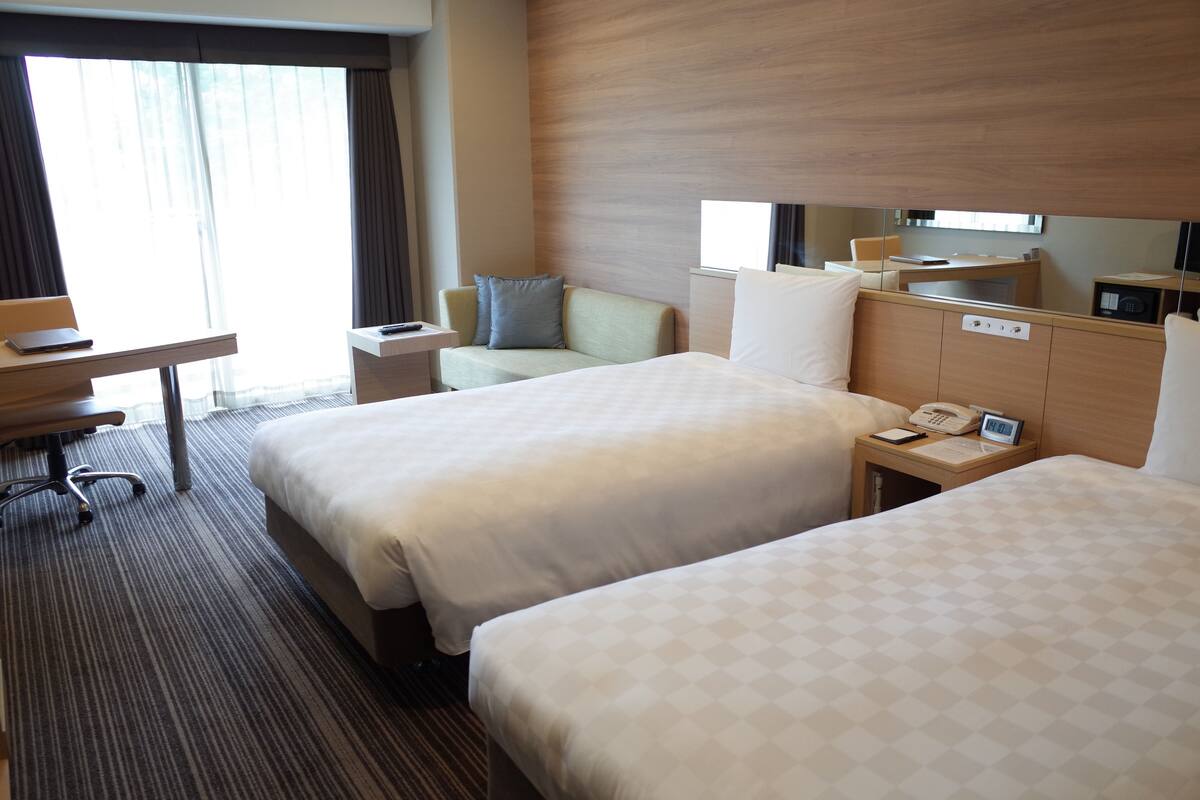 A hotel room with two beds, a desk, a chair, a bedside table with a phone, and a large window with curtains.