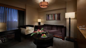 A modern living room with a sofa, armchair, coffee table with fruit and glasses, a painting on the wall, and a lamp.