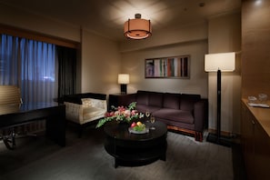 A modern living room with a sofa, armchair, coffee table with fruit and glasses, a painting on the wall, and a lamp.