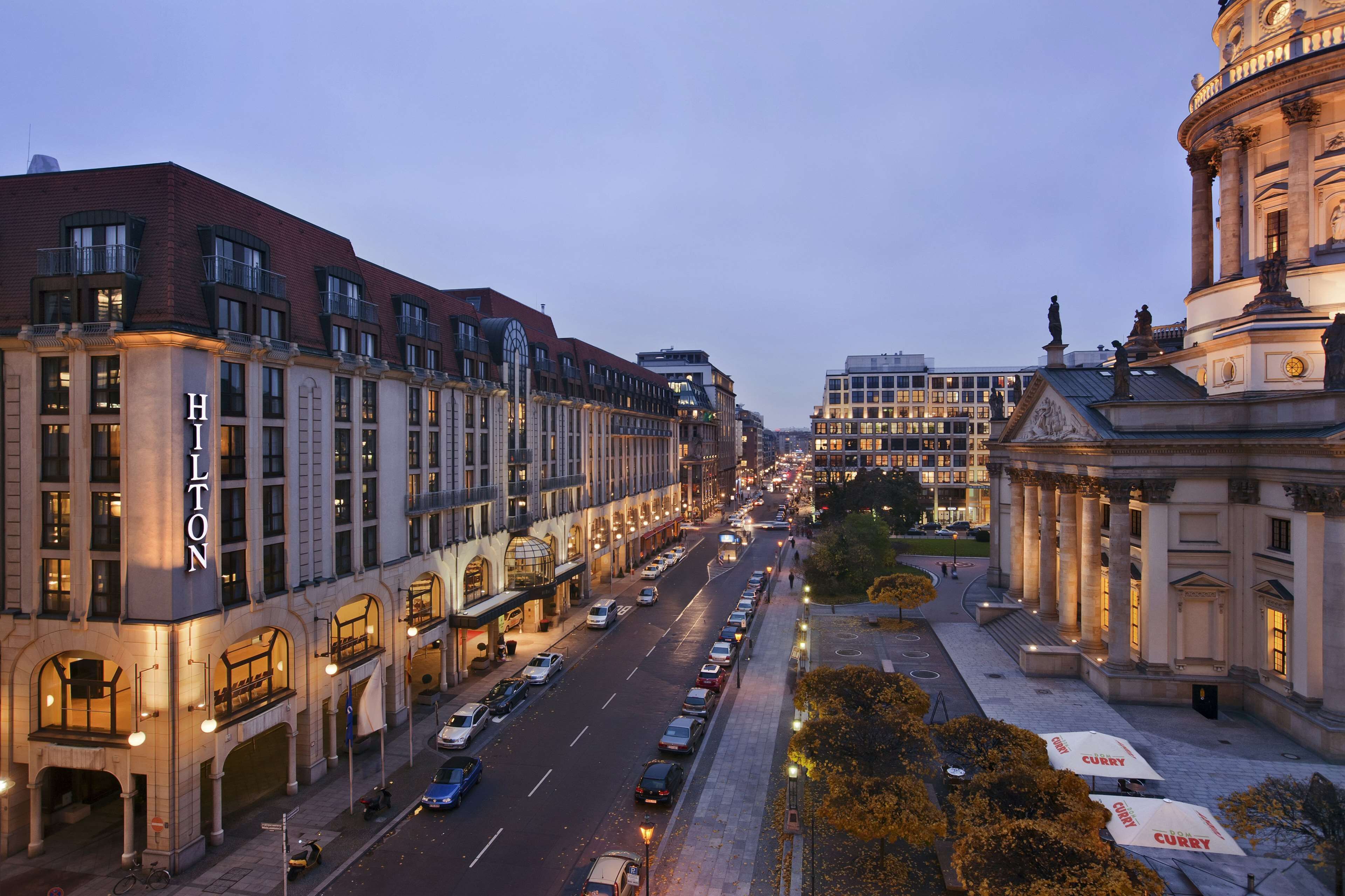 A hotel with a prominent 'Hilton' sign, a wide street with parked cars, and a classical building with columns in the background.