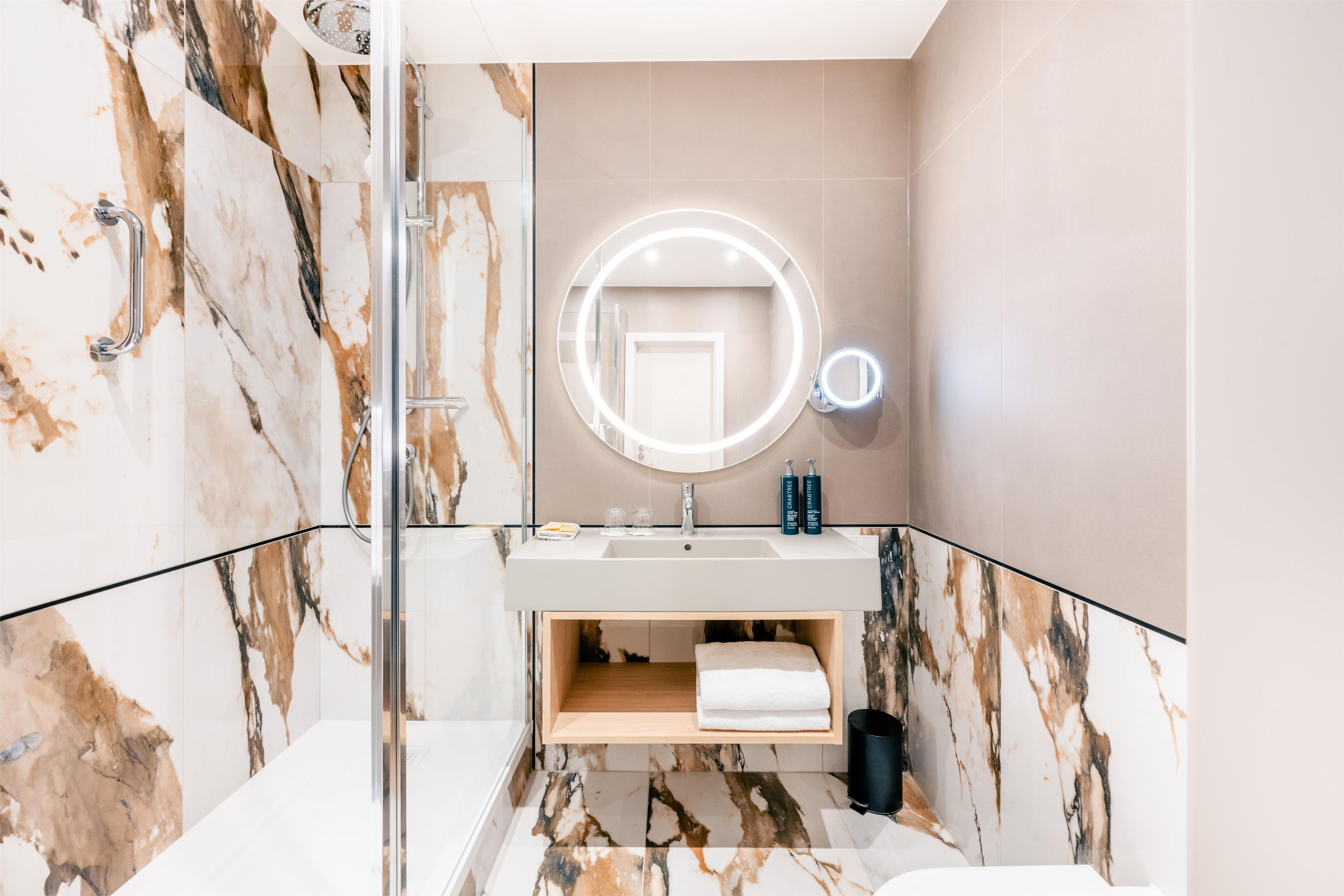 A modern bathroom with a marble vanity, a round mirror with lights, and a glass shower enclosure.