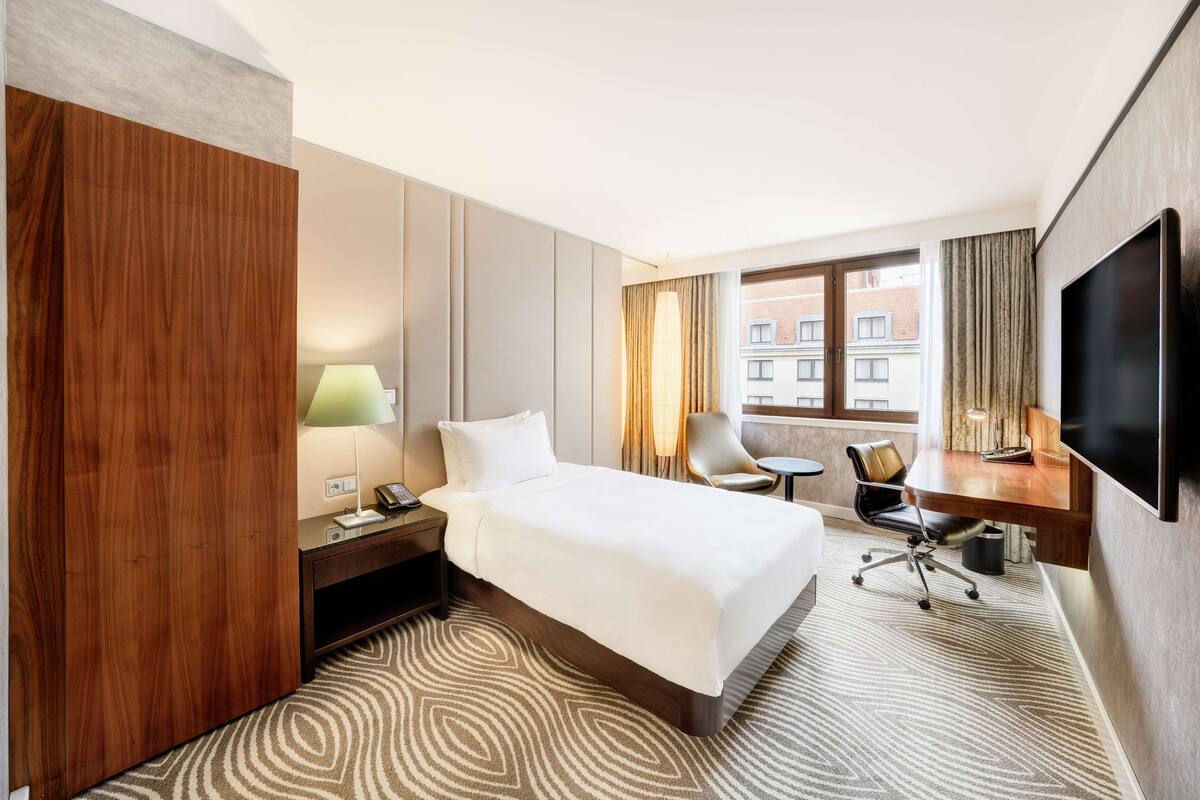 A hotel room with a large bed, a desk with a chair, a TV mounted on the wall, and a window with curtains.