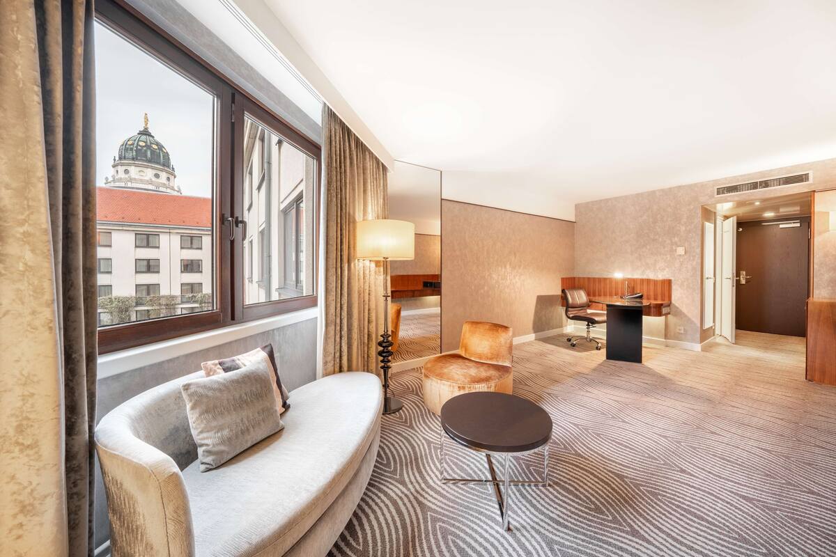 A hotel room with a sofa, a small round table, a lamp, and a view of a building with a dome through a window.