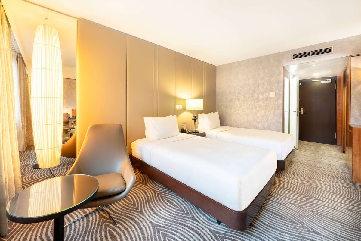A hotel room with two beds, a sitting area with a chair and a small table, a lamp, and a door leading to another room.