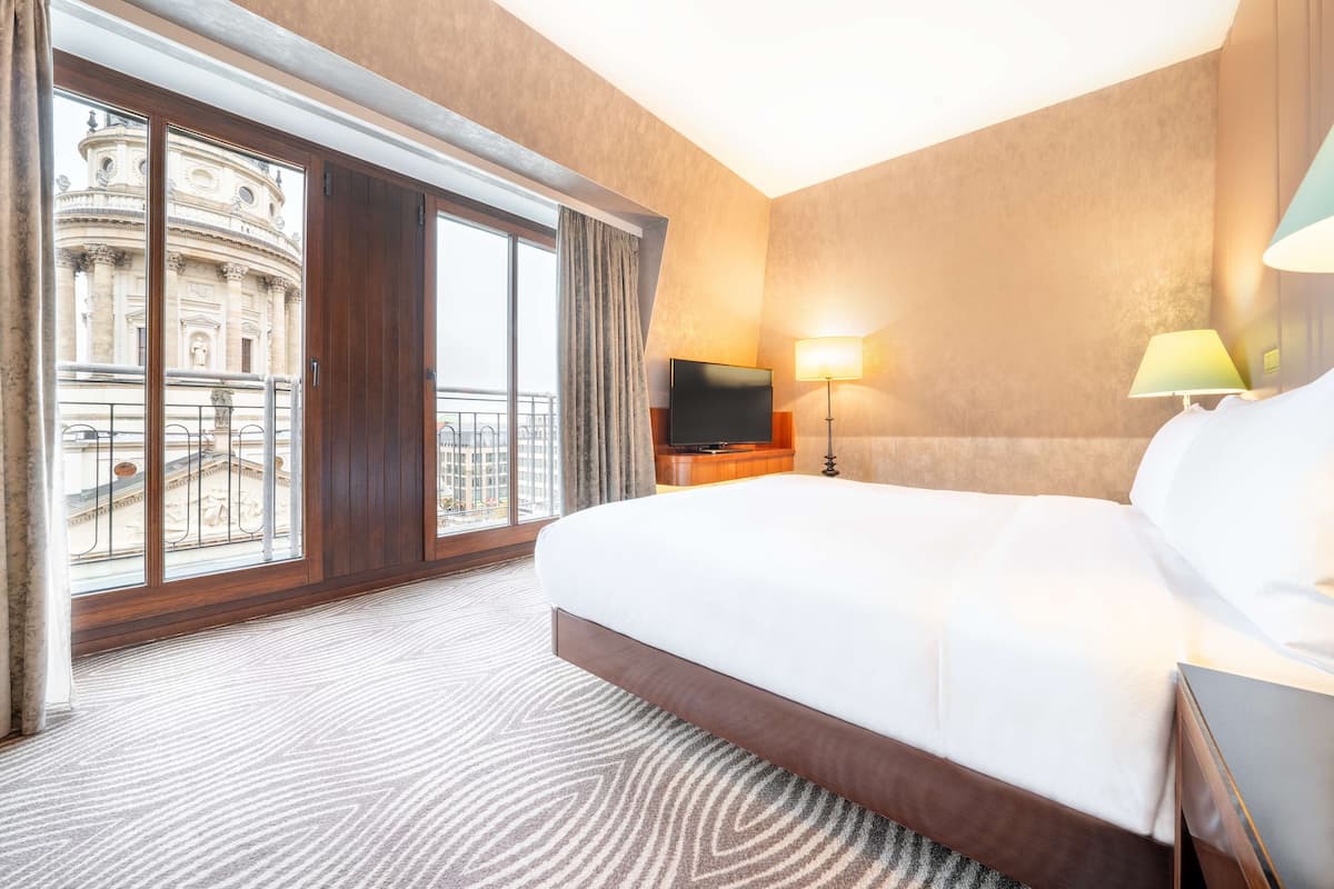 A hotel room with a large bed, a TV, a lamp, and a view of a historic building through the window.