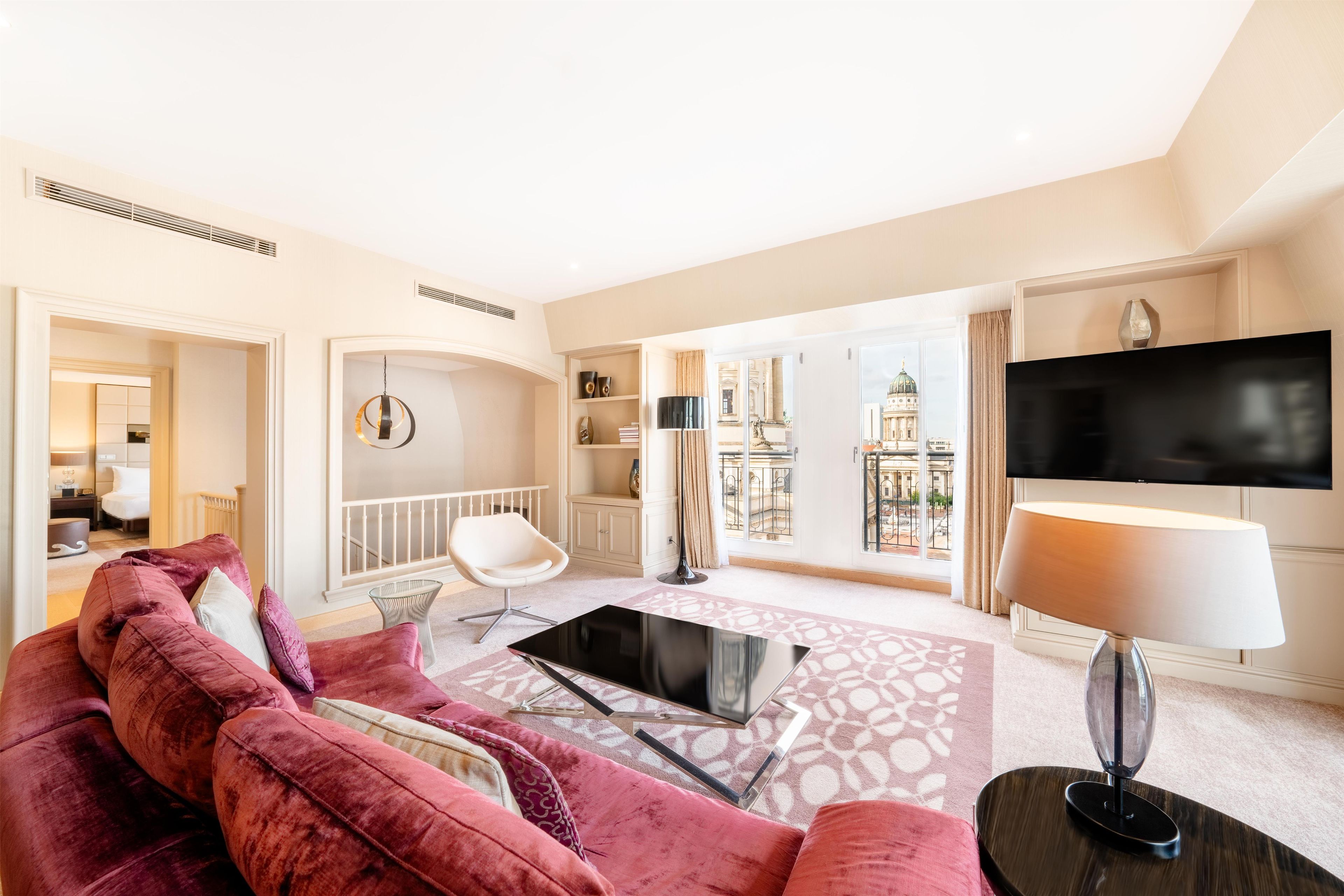 Una sala de estar moderna con un televisor de pantalla plana grande, un sofá de terciopelo rojo, una mesa de centro de vidrio y vistas de un paisaje urbano a través de ventanas amplias.