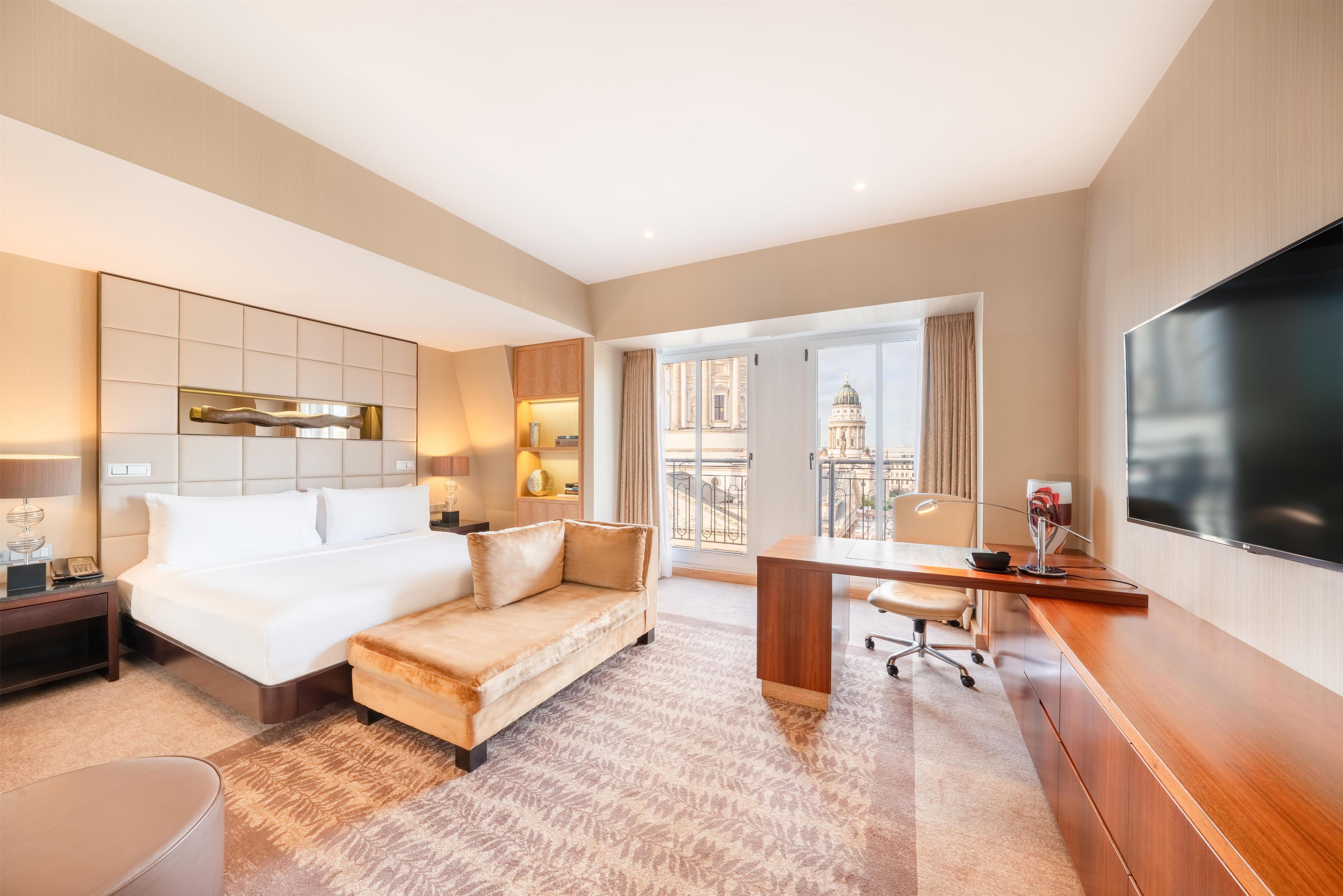 Ein Hotelzimmer mit Bett, Sofa, Schreibtisch, Fernseher und Blick auf ein Gebäude.