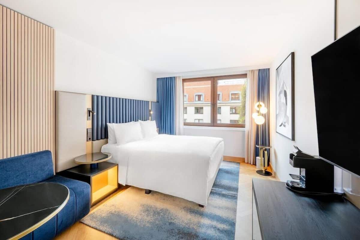 A hotel room with a bed, a desk, a chair, a TV, and a window with curtains.
