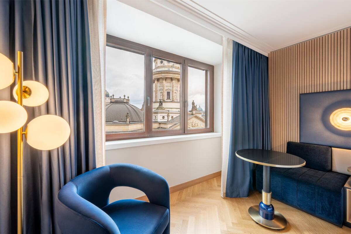 A modern hotel room with a large window, a blue chair, a round table, and a wall-mounted light fixture.