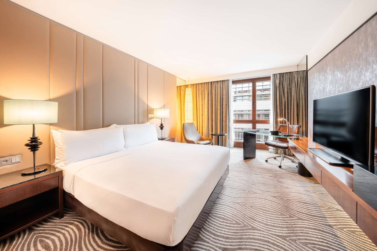 A hotel room with a large bed, a desk with a chair, a TV, and a view of the city through the window.