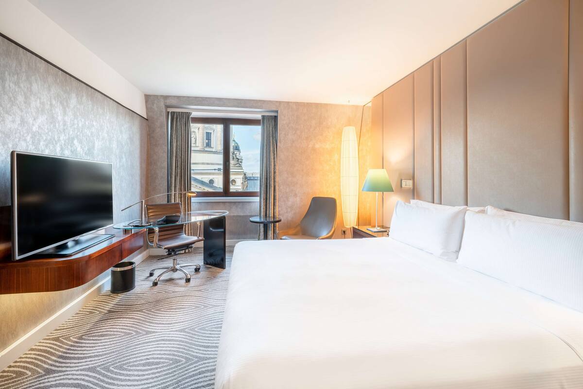 A hotel room with a large bed, a flat-screen TV, a desk with a chair, a lamp, and a view of a building through a window.