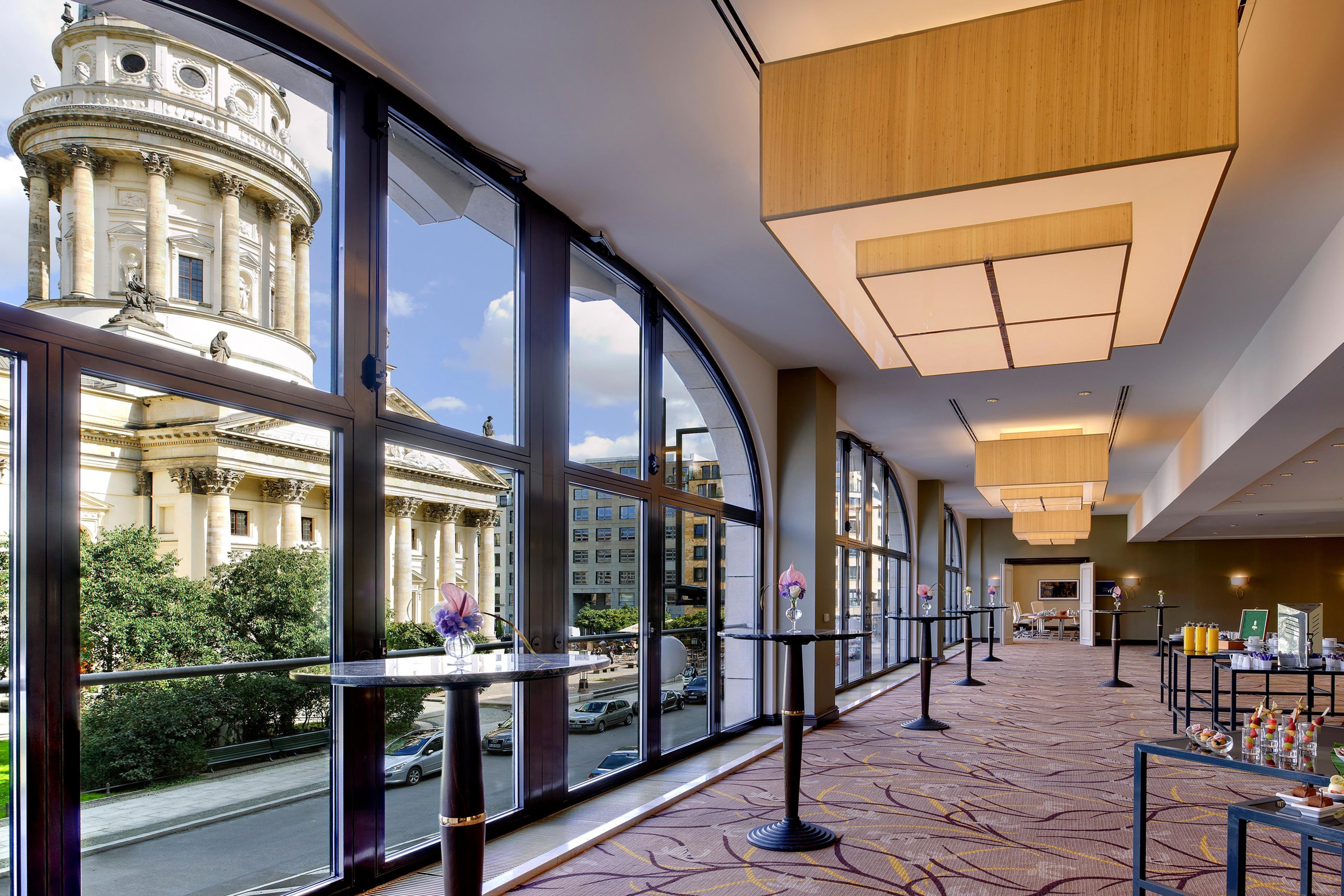 Un vestíbulo de hotel con amplias ventanas, vistas a un edificio histórico y un diseño interior moderno.