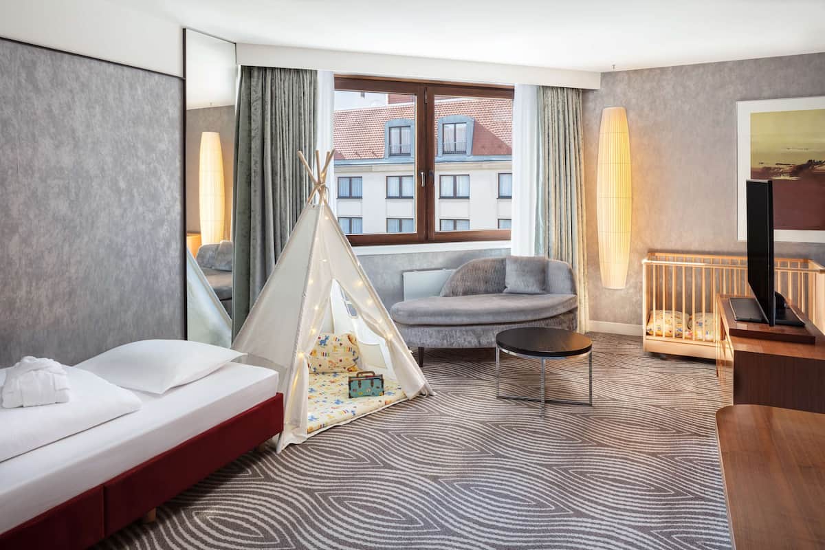 A hotel room with a bed, a sofa, a small table, a crib, a tent, and a large window with a view of buildings.