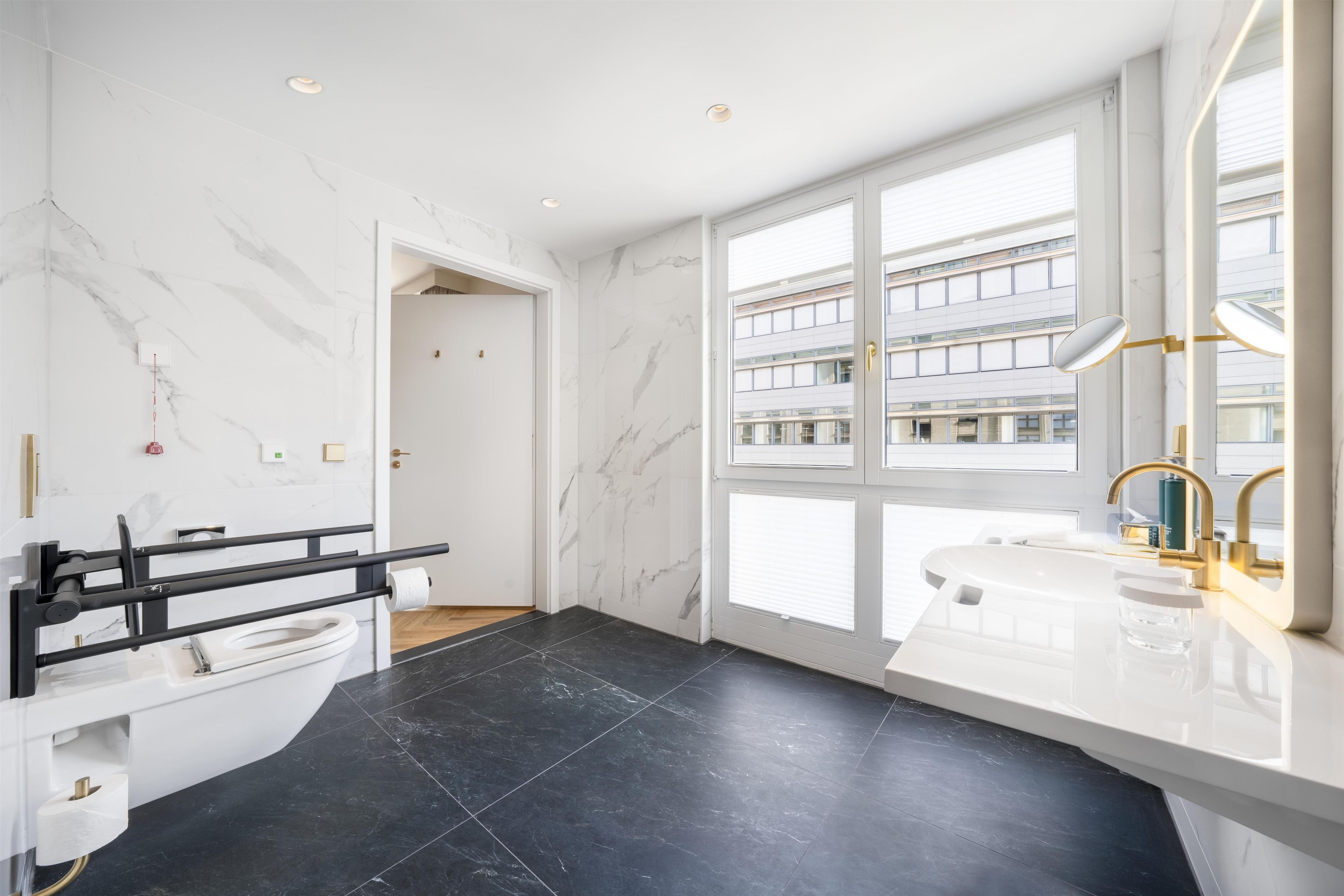 A modern bathroom with a black tiled floor, white marble walls, a large window with a view of buildings, and a gold faucet.