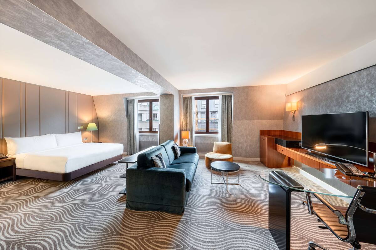 A hotel room with a bed, sofa, armchair, TV, desk, and a patterned carpet.