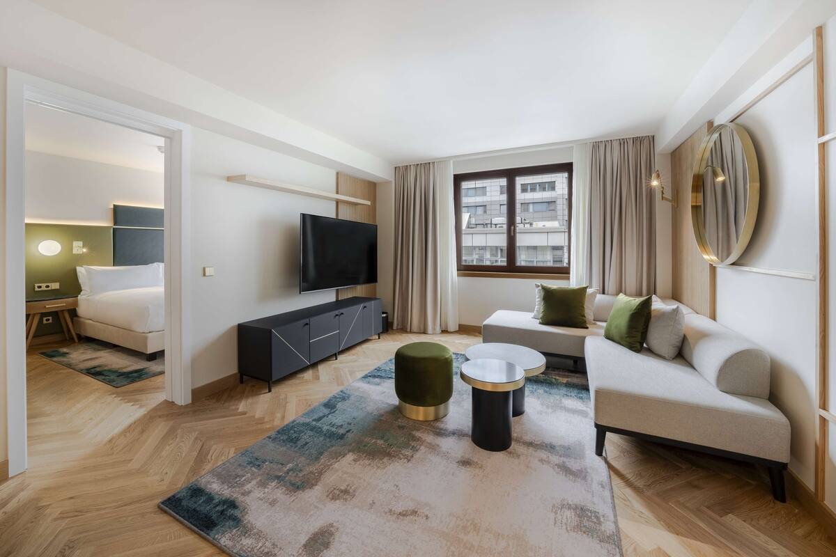 A modern hotel room with a sofa, a TV, a small table, and a large window.