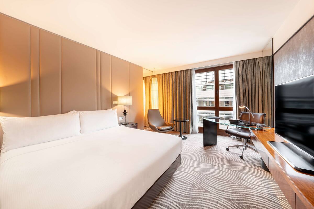 A hotel room with a large bed, a desk with a chair, a television, and a window with curtains.