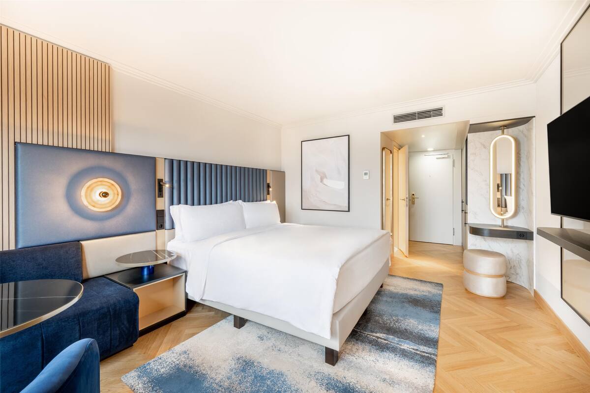A modern hotel room with a large bed, a seating area with a table, a TV, and a bathroom with a mirror and shower.