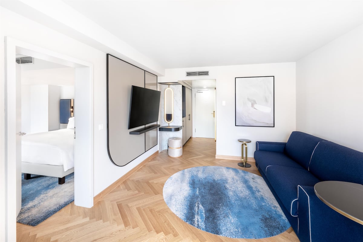 A modern hotel room with a bed, a blue sofa, a round coffee table, a TV, and artwork on the walls.