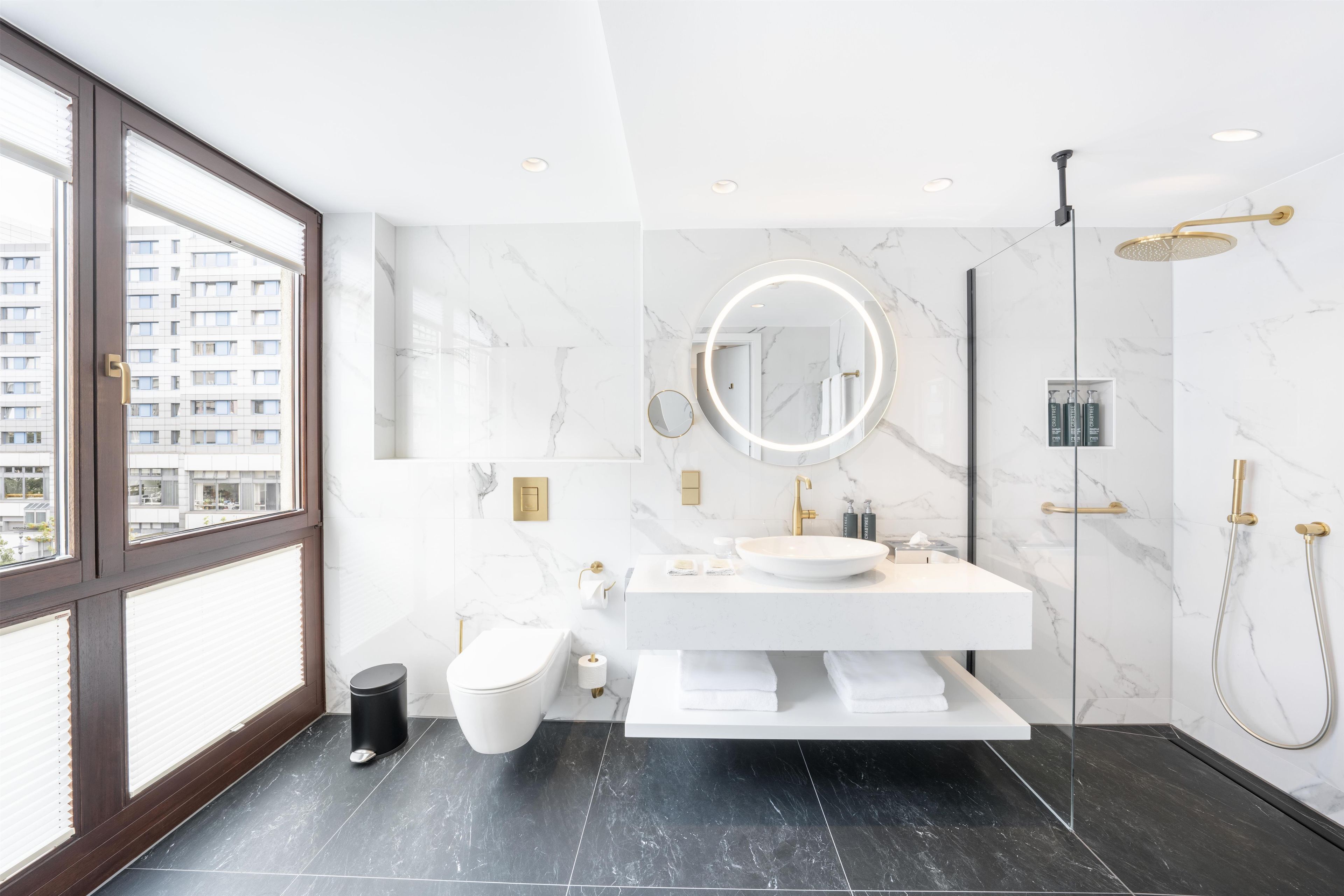 A modern bathroom with a large window, a round mirror, a white sink, a toilet, and a shower area.