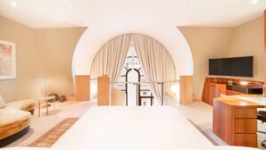 A modern hotel room with a large bed, a desk, a TV, and a view through an arched window.