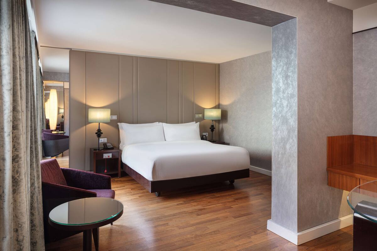 A hotel room with a bed, bedside tables, a chair, and a glass table.