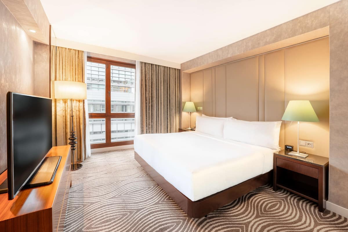 A hotel room with a large bed, a flat-screen TV, two bedside lamps, a nightstand, and a view of the city through large windows.