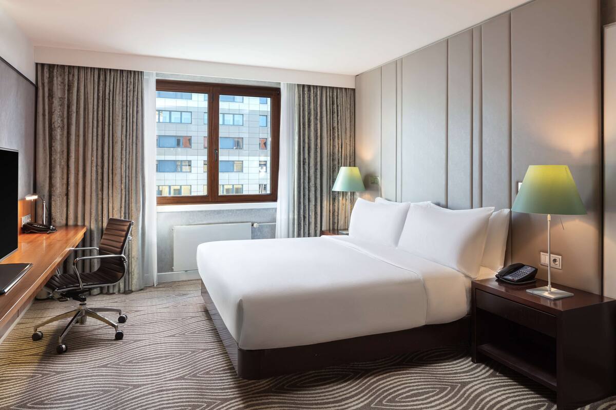 A hotel room with a large bed, a desk with a chair, a TV, two lamps, and a view of buildings through the window.