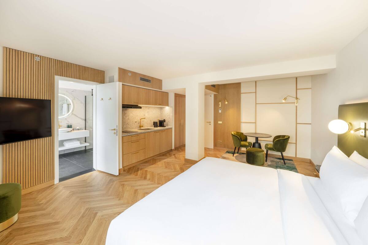 A modern hotel room with a large bed, a TV, a kitchenette, and a bathroom.