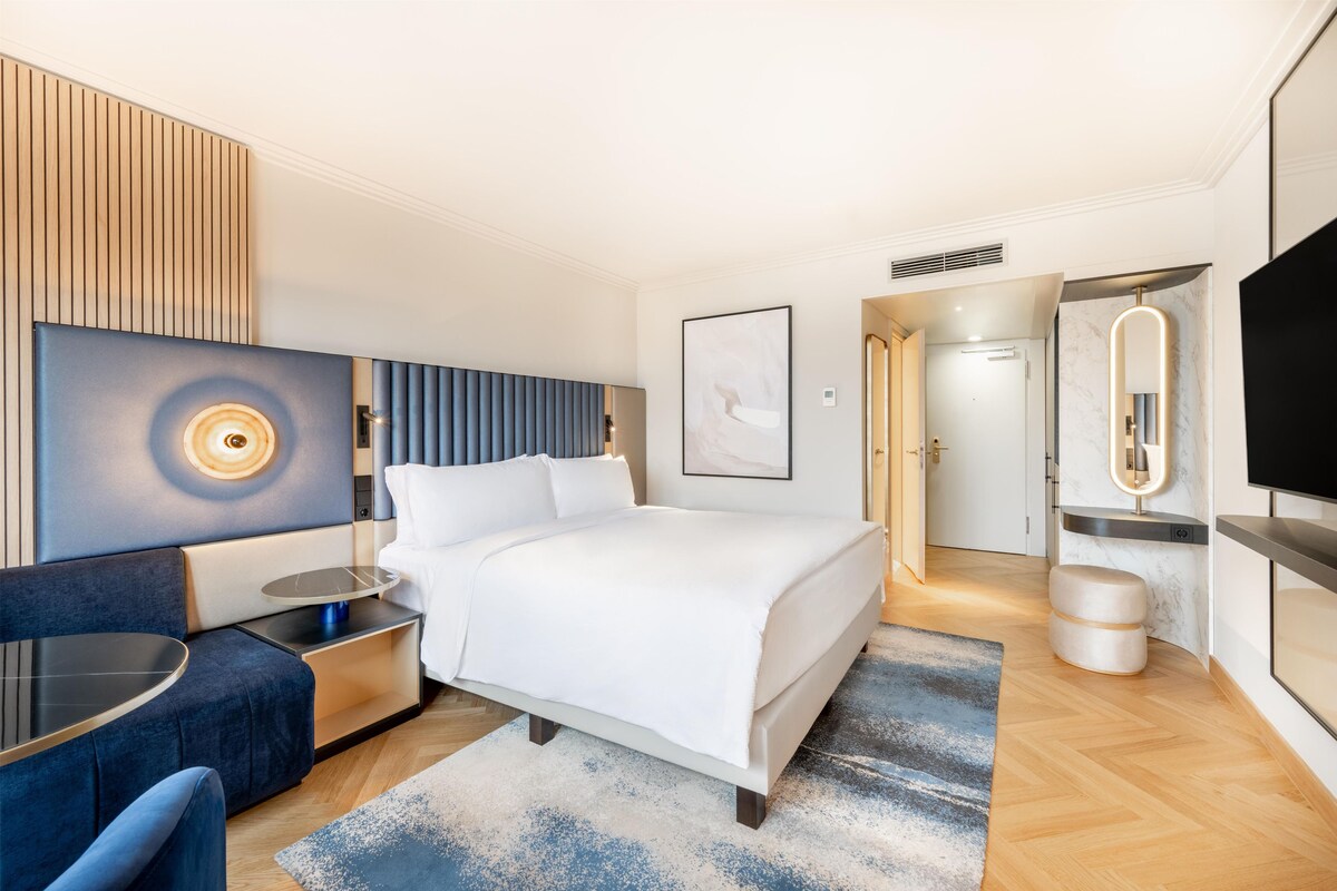 Quarto de hotel moderno com cama, mesa de cabeceira, área de estar e televisão.