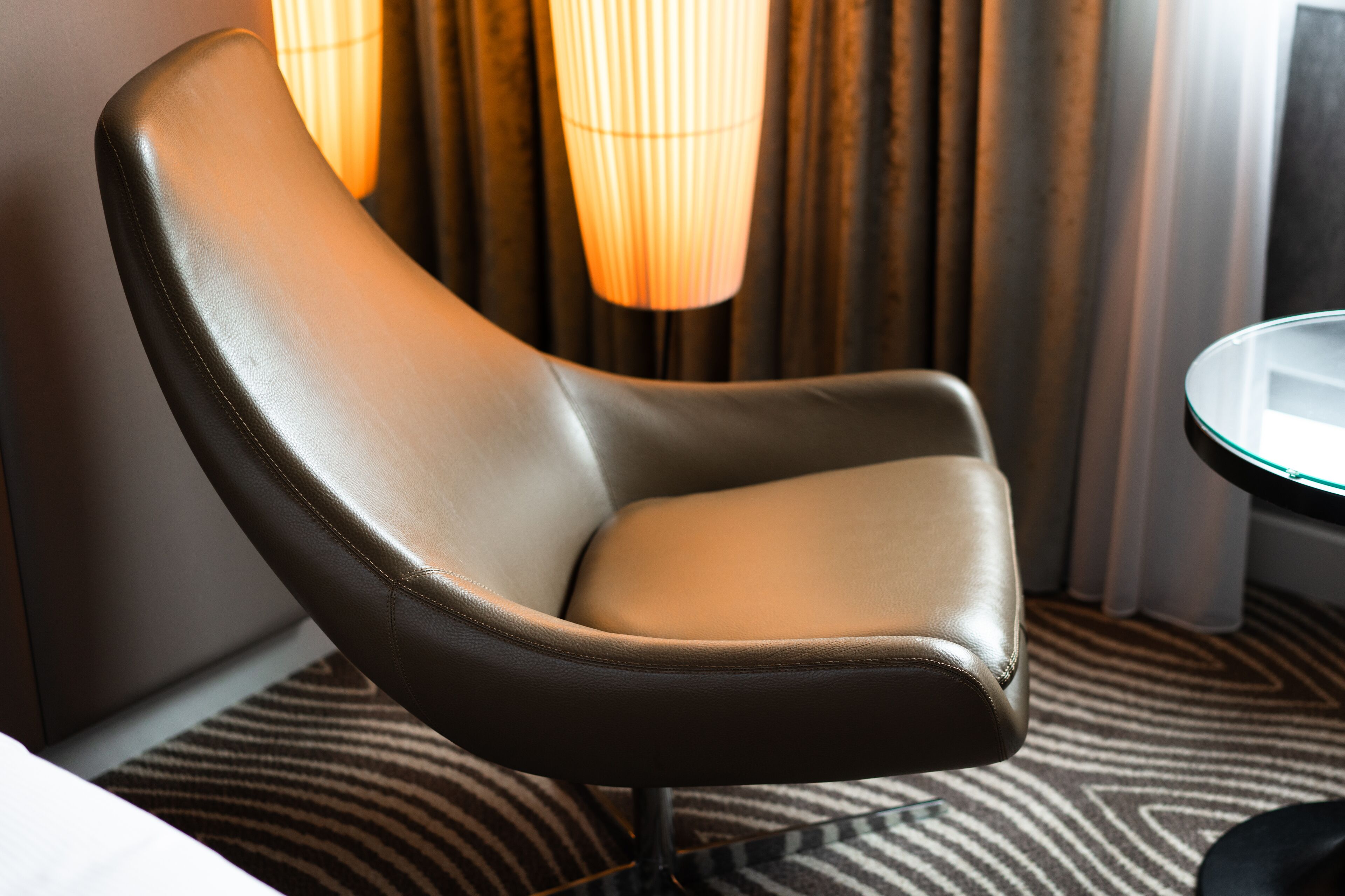 A modern chair with a curved backrest and a cushioned seat, positioned in a room with a patterned carpet and a lamp in the background.