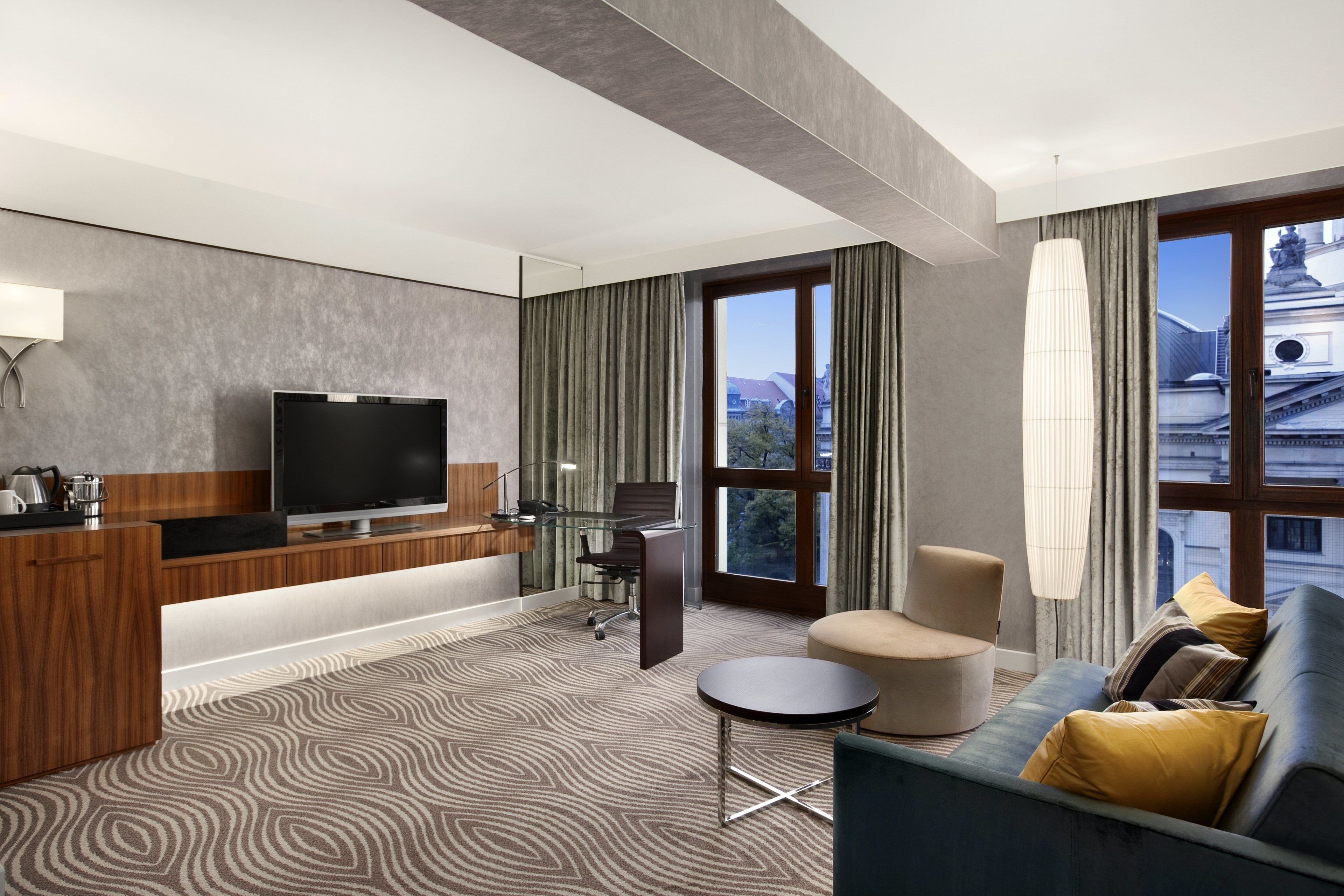 Une chambre d’hôtel moderne dotée d’une télévision à écran plat, d’un bureau avec une lampe, d’un canapé et d’une grande fenêtre donnant sur la ville.