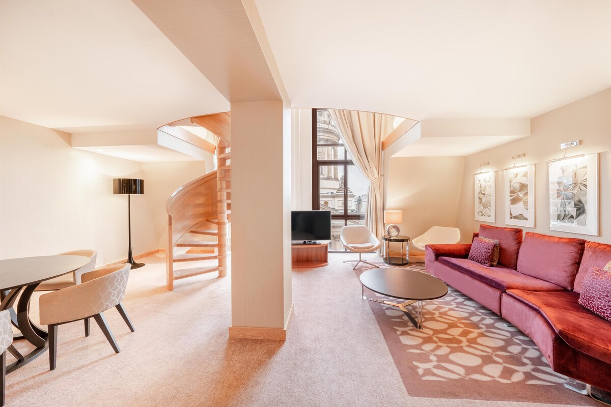 Quarto de hotel moderno com sofá, mesa de jantar, cadeiras, TV e escada para um andar superior.