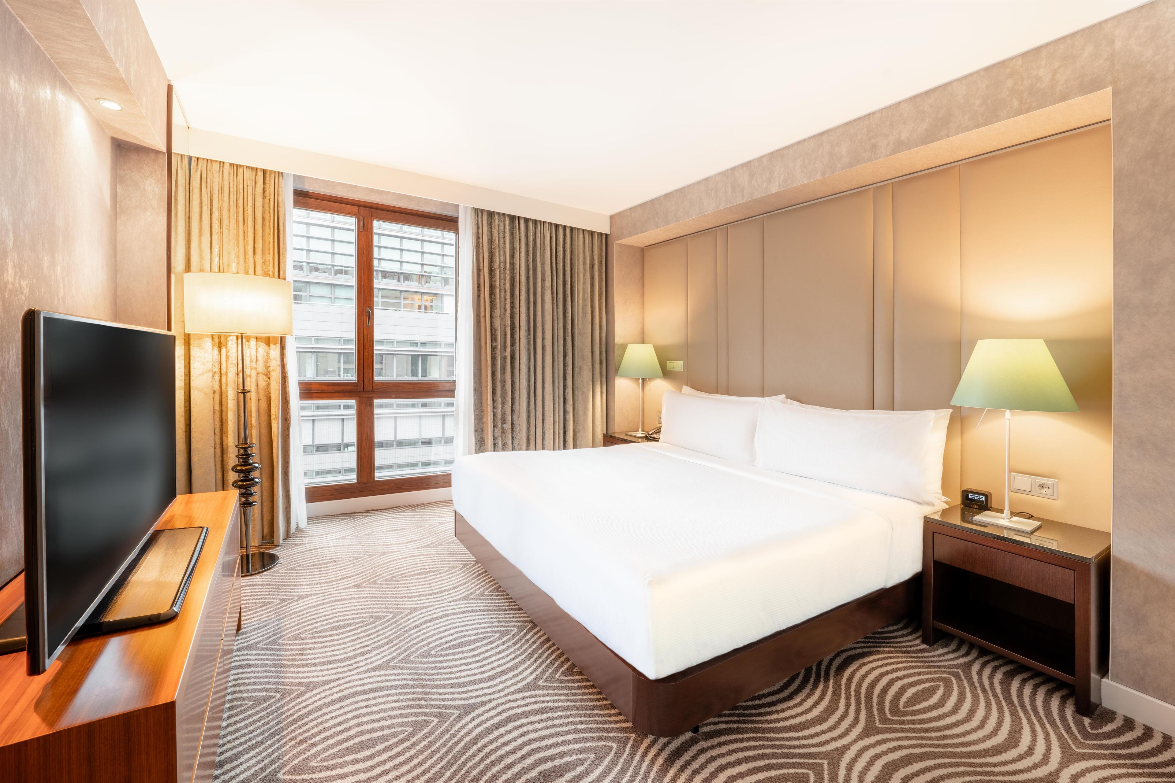 A hotel room with a bed, a TV, two bedside lamps, a window with curtains, and a patterned carpet.